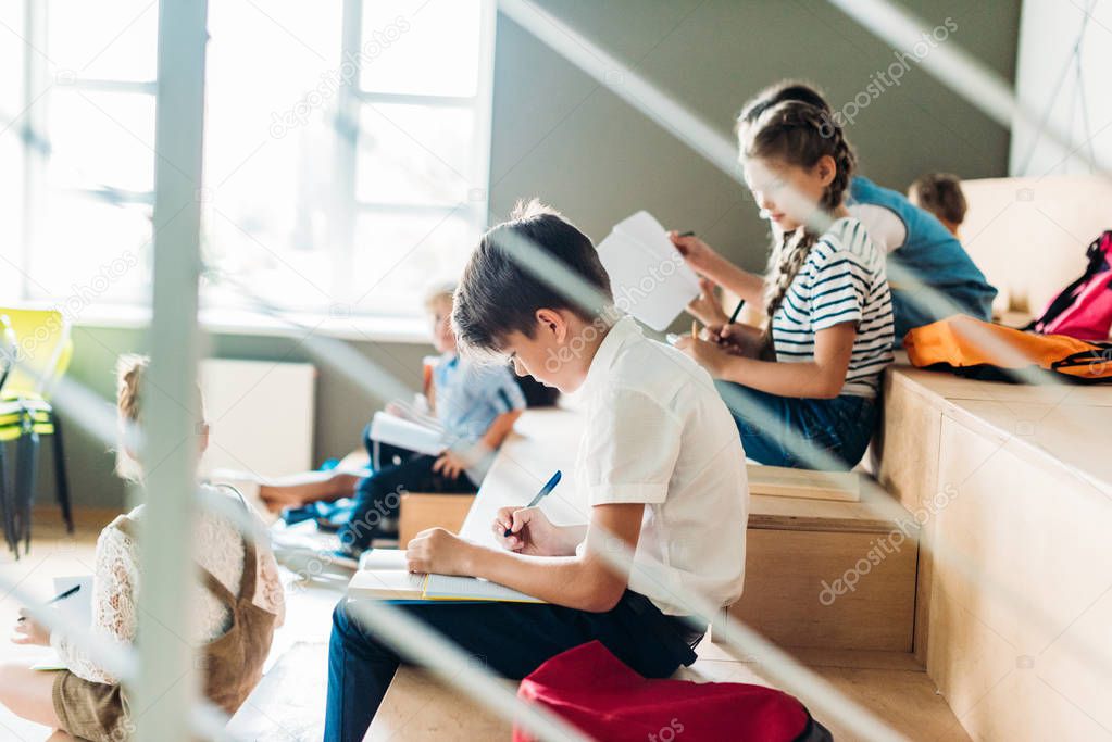 side view of young schoolboy writing in notebook while sitting on tribune at school with classmates