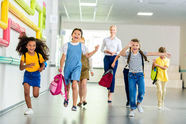 очаровательные счастливые школьники, бегущие по школьному коридору вместе с учителем, идущим позади
