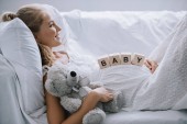 side view of smiling pregnant woman in white nightie with teddy bear and wooden blocks with baby lettering on belly resting on sofa