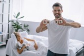 selective focus of smiling man showing wooden blocks with baby inscription while pregnant woman resting on sofa