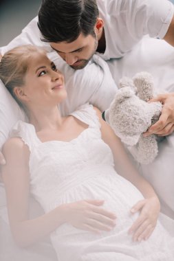 oyuncak ayı ve evde kanepe üzerinde beyaz Geceliği gülümseyen hamile karısı olan adam portresi