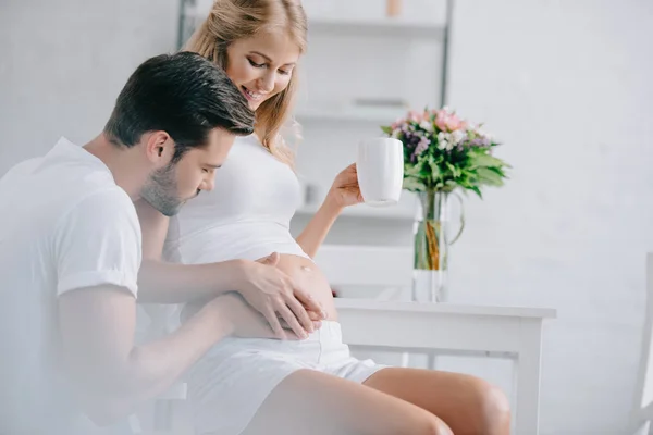 Sonriente Mujer Embarazada Con Taza Descansando Silla Mientras Marido Tocando —  Fotos de Stock