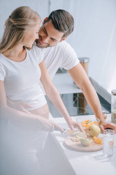happy pregnant woman and husband cooking fruits salad together in kitchen at home