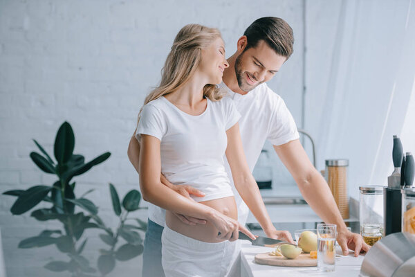 happy pregnant woman and husband cooking fruits salad together in kitchen at home