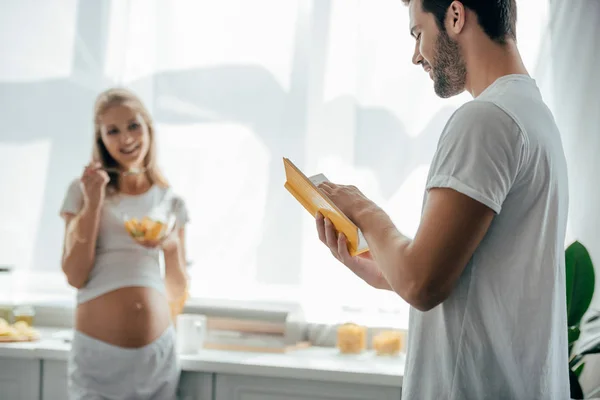 フルーツ サラダと妊娠中の妻と夫の自宅の台所での本の選択と集中  — 無料ストックフォト