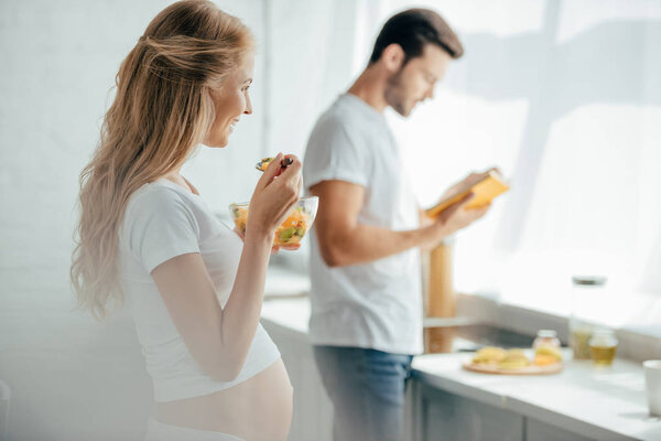 selective focus of pregnant woman eating fruits salad while husband with book standing at counter in kitchen