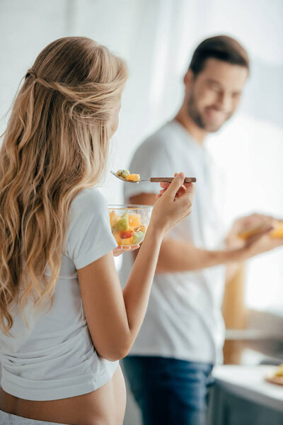 selective focus of pregnant woman eating fruits salad while smiling husband standing at counter in kitchen