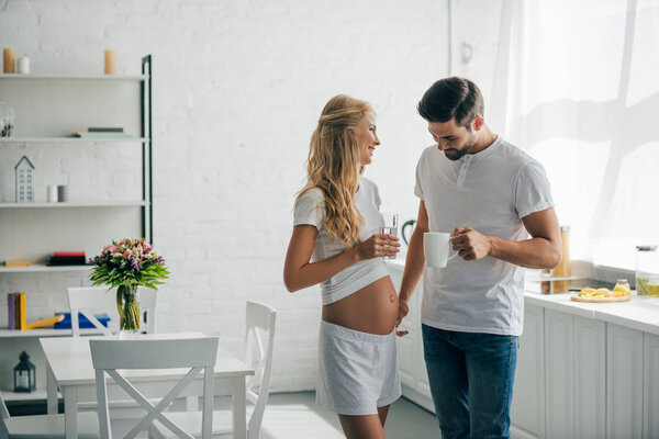man with cup of coffee looking at belly of pregnant wife in kitchen at home
