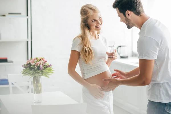 家庭での水のガラスと陽気な妊娠中の妻の腹に触れる夫 — ストック写真
