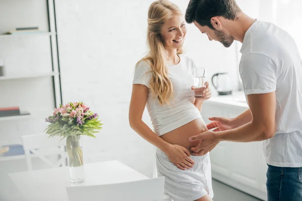 家庭での水のガラスで幸せな妊娠中の妻の腹に触れる夫 — ストック写真