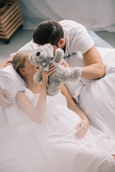 模糊的人和怀孕的妻子在白色睡衣持有泰迪熊在家沙发上 — 图库照片