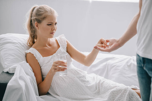 partial view of man giving medicines to pregnant woman in white nightie on sofa at home