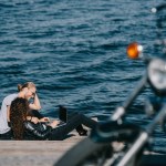 Young couple using laptop while sitting on quay near the sea, selective focus of motorcycle