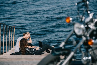 Genç çift iskele üzerinde otururken dizüstü kullanarak yakınındaki deniz, motosiklet seçici odak