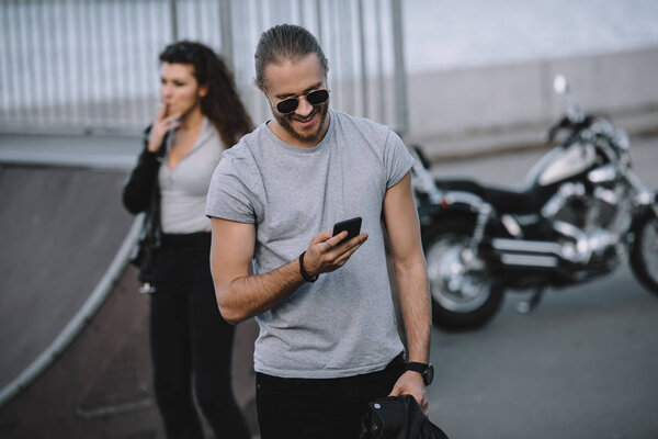 man using smartphone while girlfriend standing on background with classic motorbike 