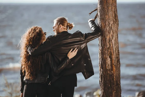 back view of couple in black leather jackets hugging and looking at each other on seashore