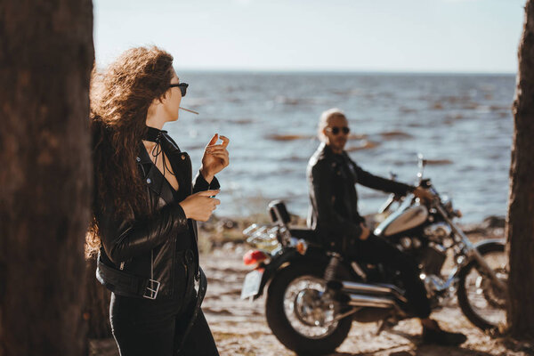 selective focus of woman smoking cigarette and looking at her boyfriend on motorbike on seashore