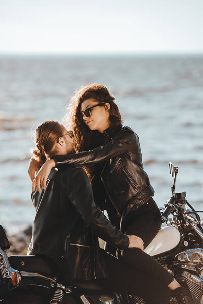 couple of bikers in black leather jackets embracing on classic motorcycle on seashore