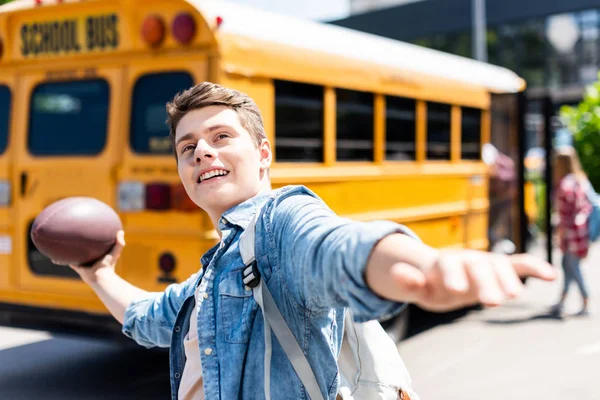 Счастливый Школьник Бросает Американский Футбольный Мяч Перед Школьным Автобусом — стоковое фото