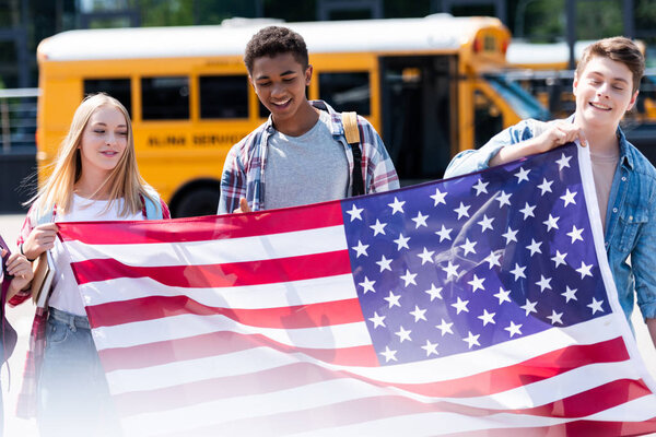группа улыбающихся подростков, держащих флаг США перед школьным автобусом
