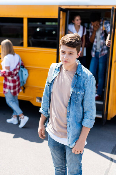 depressed teenager schoolboy standing in front of school bus with classmates