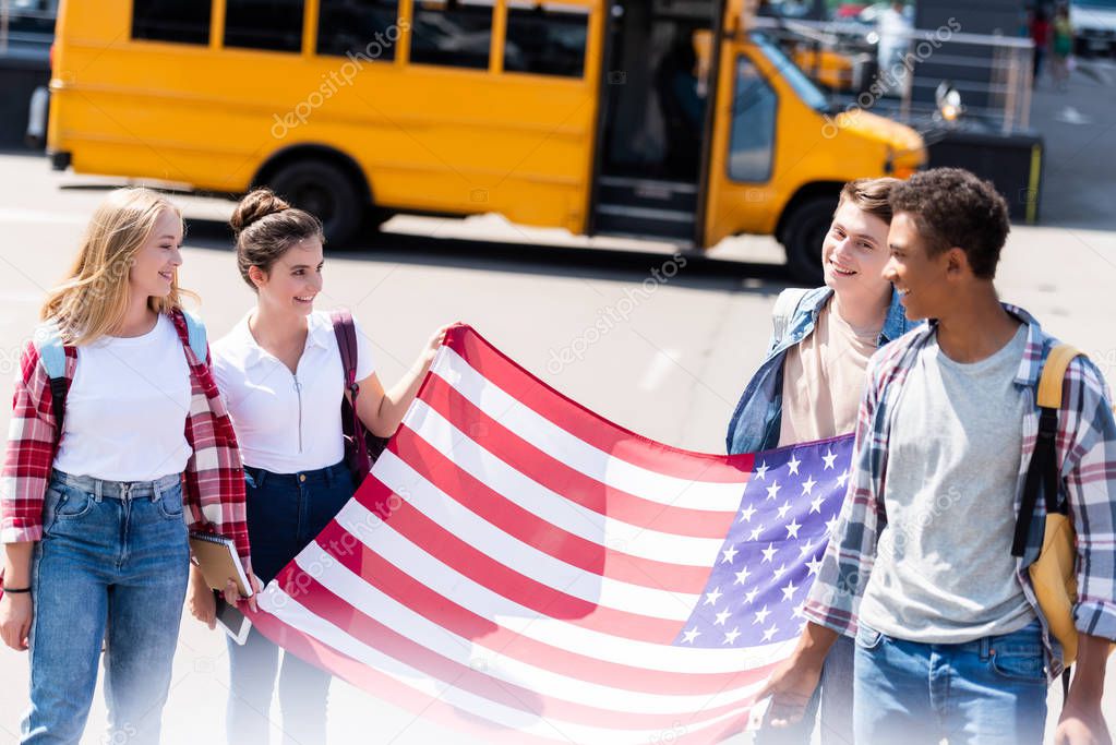 group of multiethnic american teen scholars with usa flag in front of school bus