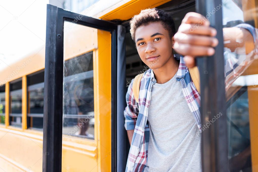 close-up portrait of happy teen african american schoolboy standing in door of school bus and looking at camera