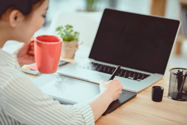 Genç Asyalı tasarımcı grafik tablet ve dizüstü bilgisayar ile çalışırken kahve içme