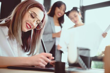 young designer drawing on graphics tablet while her colleagues looking at blueprint clipart