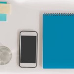 Pohled shora na smartphone s prázdnou obrazovkou, zápisník s perem, sklenice vody a poznámek sticky notes