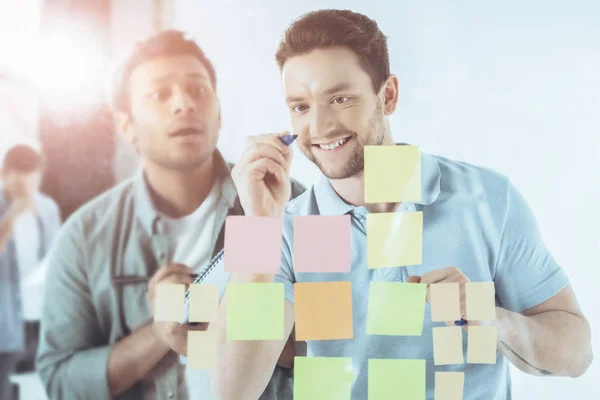 Smiling Young Businessman Writing Sticky Notes While Shocked Colleague Standing — Free Stock Photo