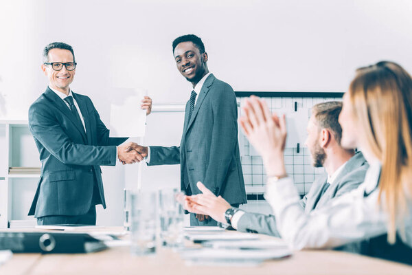 Кавказский босс пожимает руку африканскому американскому менеджеру, пока коллеги хлопают в ладоши
