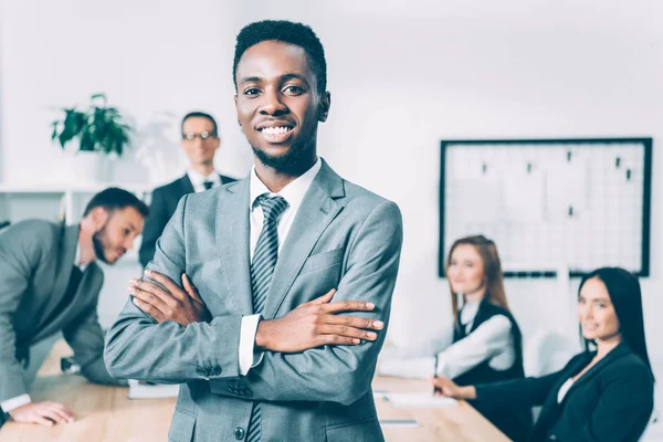 handsome african american manager with crossed arms with blurred multicultural colleagues on background