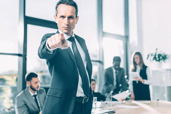 Ernstige Zakenman Wijzend Camera Conferentiezaal Met Wazig Collega Achtergrond — Gratis stockfoto