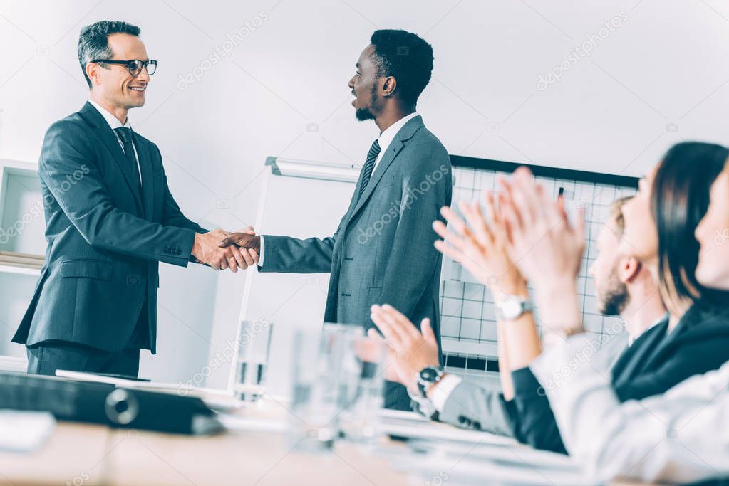 multiethnic businessmen shaking hands in conference hall with partners sitting at table and claping hands