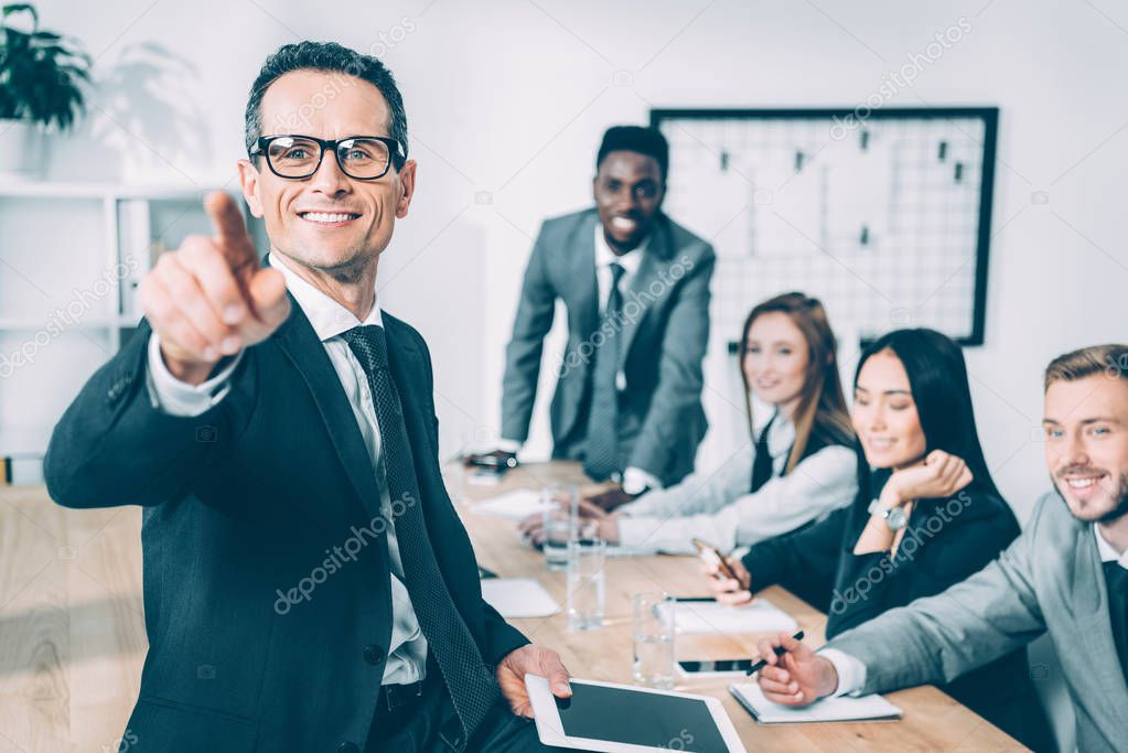 handsome businessman pointing at camera with multicultural partners sitting in conference hall blurred on background