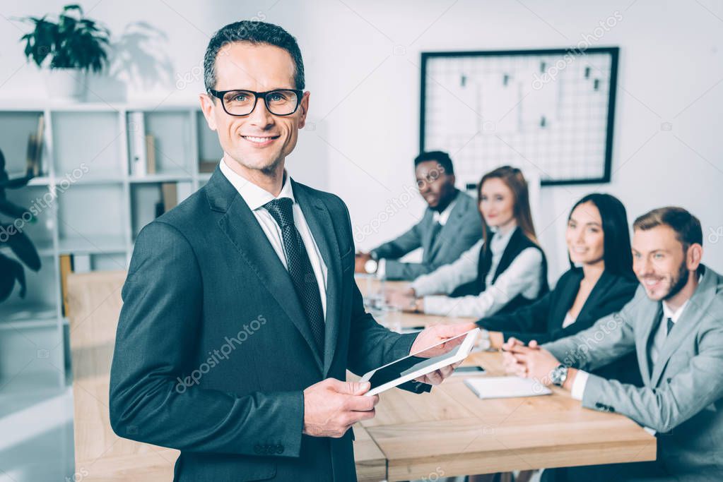 handsome businessman holding tablet in conference hall with multicultural partners sitting at table