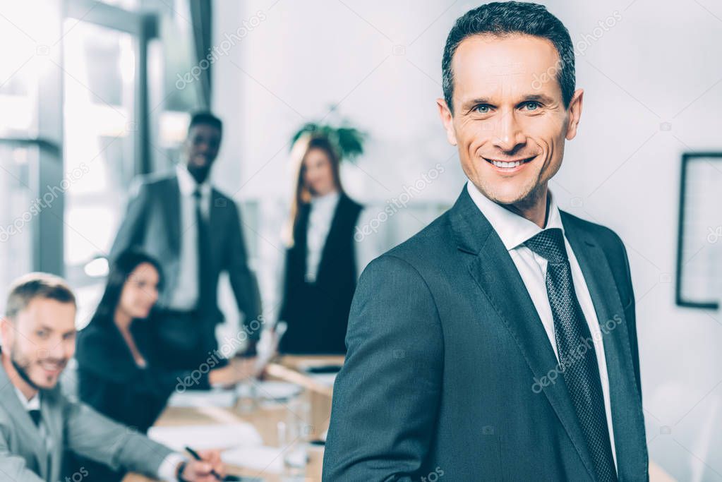 handsome businessman looking at camera in conference hall with multiracial partners blurred on background