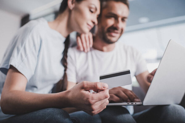 селективный фокус улыбающейся пары с ноутбуком, делающей покупки по кредитной карте дома
