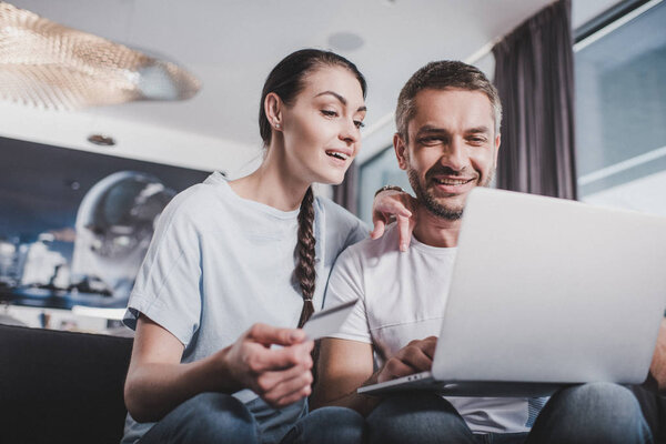 улыбающаяся пара с ноутбуком, делающая покупки по кредитной карте дома
