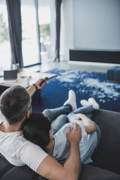 high angle view of adult man with remote controller embracing girlfriend and watching tv on sofa at home