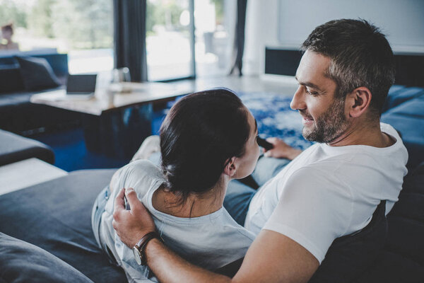 улыбающийся мужчина с пультом дистанционного управления обнимает подругу и смотрит телевизор на диване дома
