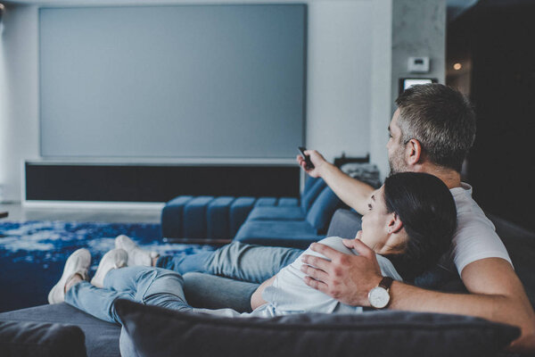 adult man with remote controller embracing girlfriend and watching tv on sofa at home