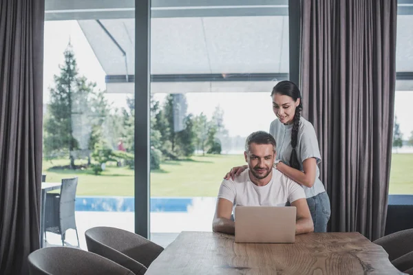 Sonriente Joven Mujer Abrazando Novio Mientras Usando Ordenador Portátil Casa — Foto de stock gratuita