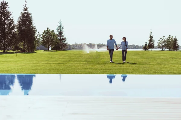 夫妇牵着手 走在绿色的草坪附近的游泳池在乡间别墅 — 免费的图库照片