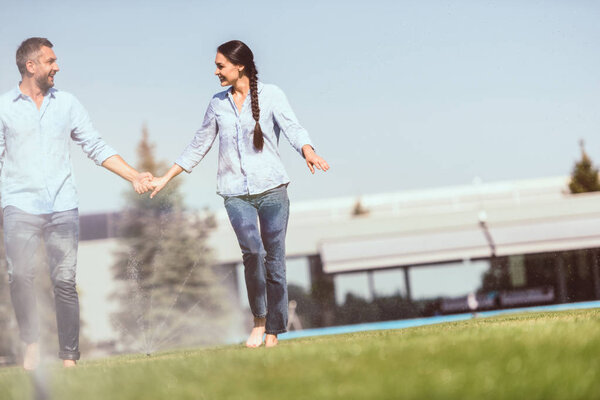 happy couple holding hands and having fun with automatic watering on grass near country house 