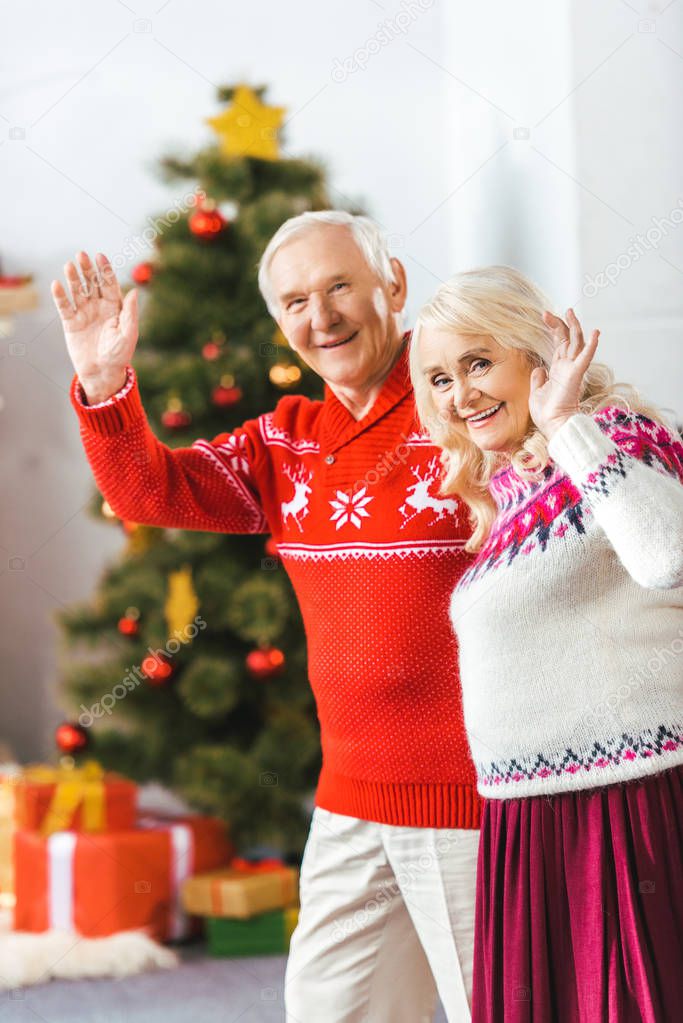 happy senior couple in christmas sweaters waving at camera