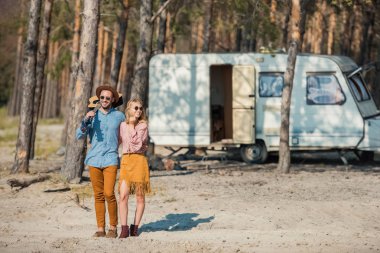 young hippie couple hugging and standing with guitar near campervan in forest clipart