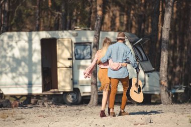 arkadan sarılma ve kamp aracına yakınındaki akustik gitar ile yürüme hippi çift görünüşü