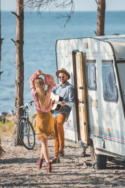 hippie couple dancing and playing guitar near trailer in nature